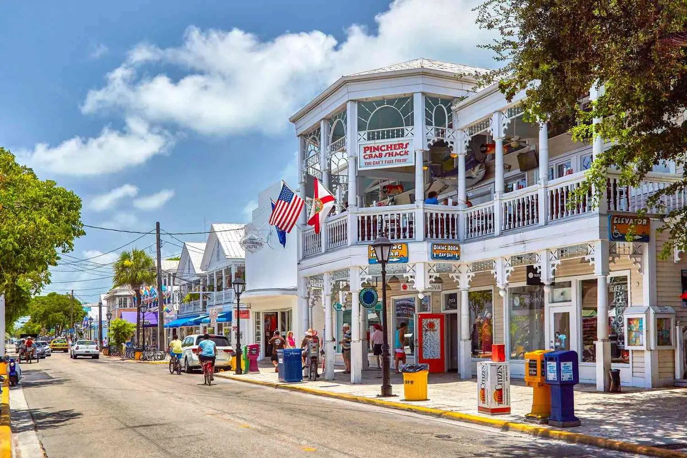 A street scene with a white building, people, cars, and trees under a blue sky with clouds.