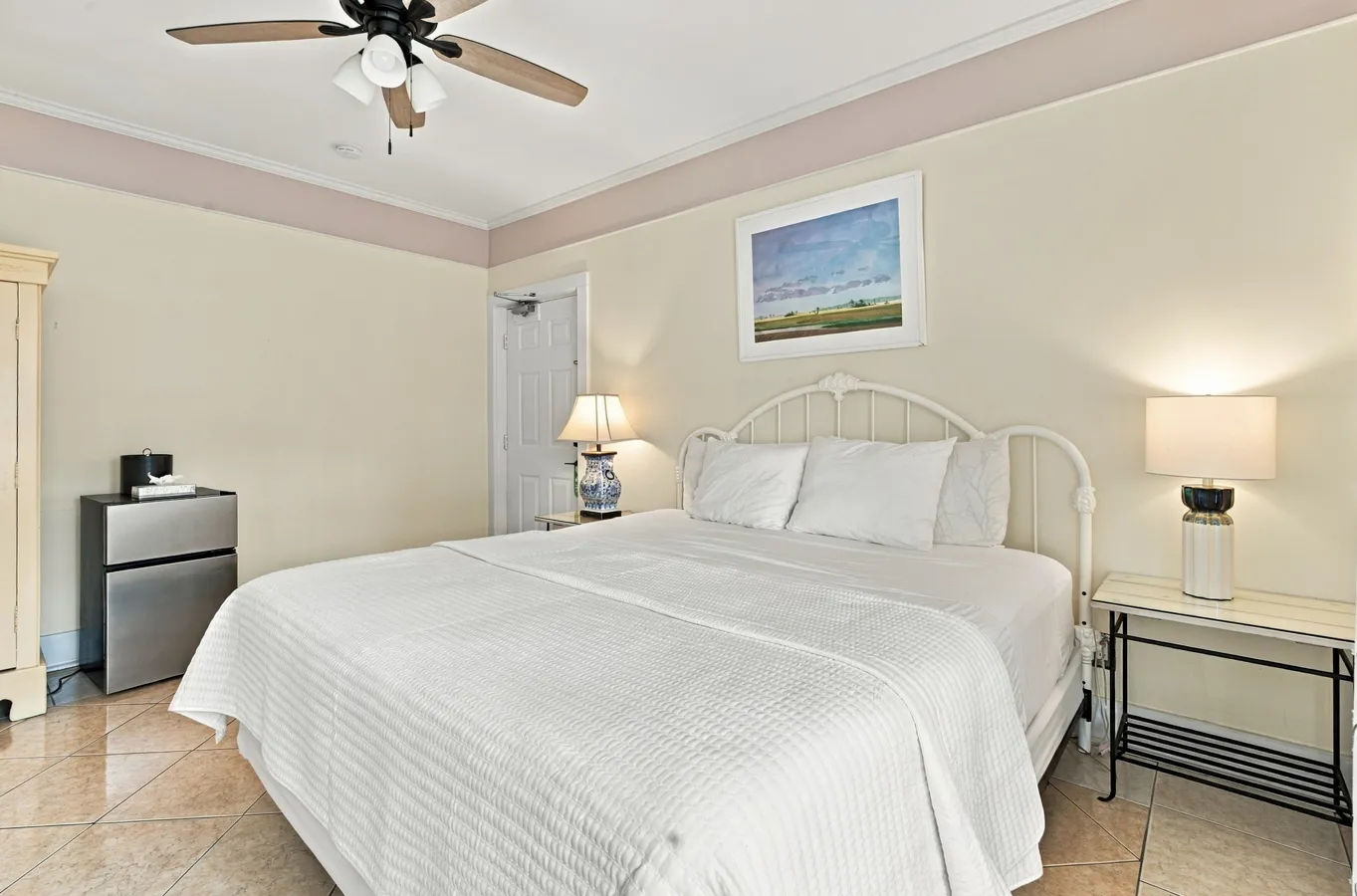 A hotel room with a white bed, nightstand, mini fridge, and ceiling fan.